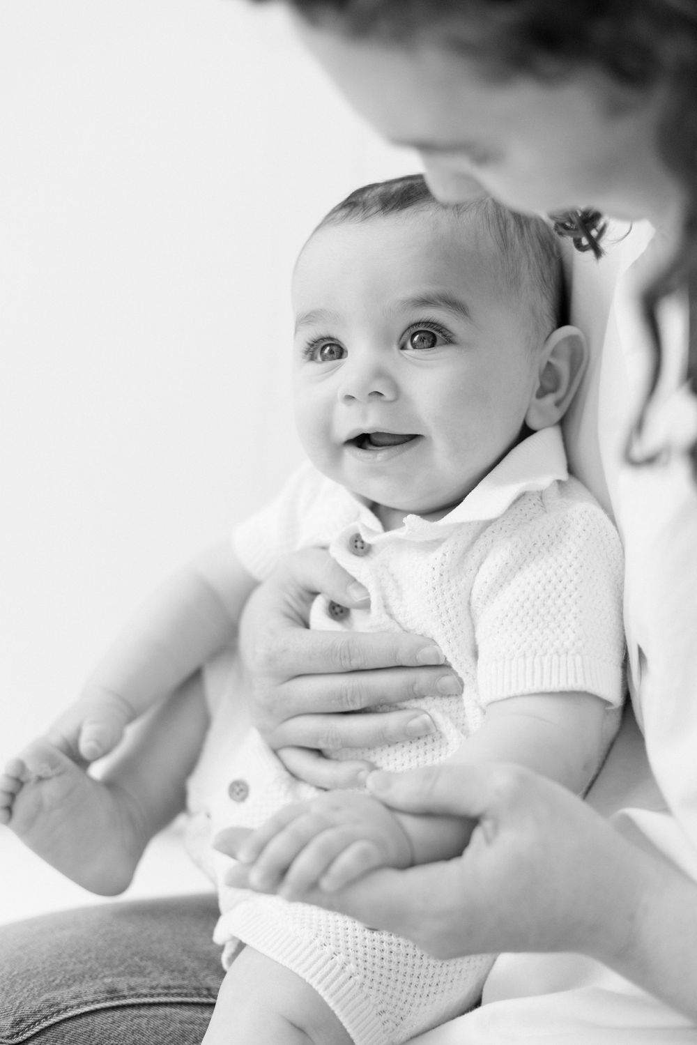 A black and white portrait of a smiling baby in a white knit outfit being held by gentle hands.