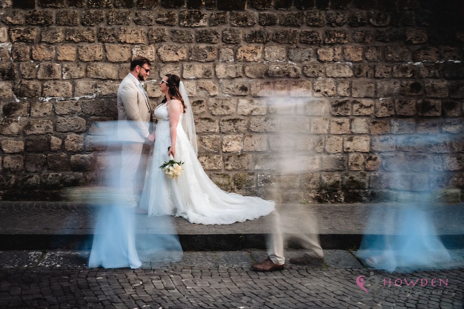 Artistic wedding photo against brick wall with motion blur showing walking people in blue passing by.