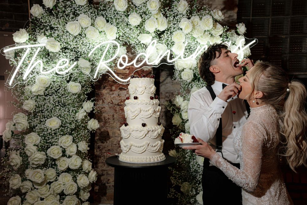 Wedding cake cutting moment under floral arch with neon sign.