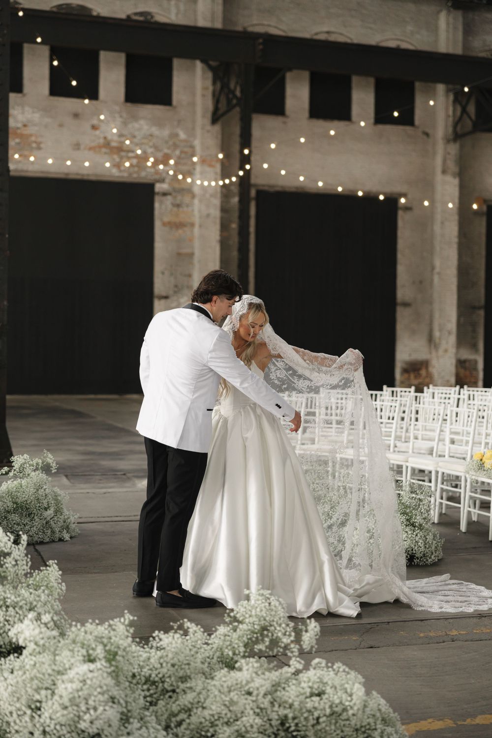 Romantic moment between newlyweds in industrial venue with string lights and white chairs.