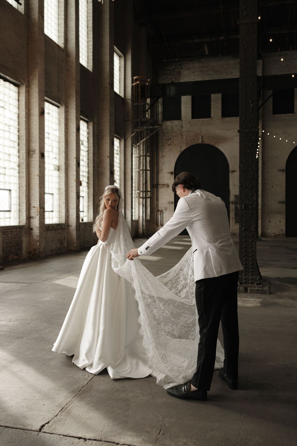 A bride in a white gown has her dress adjusted in an industrial venue with large windows.