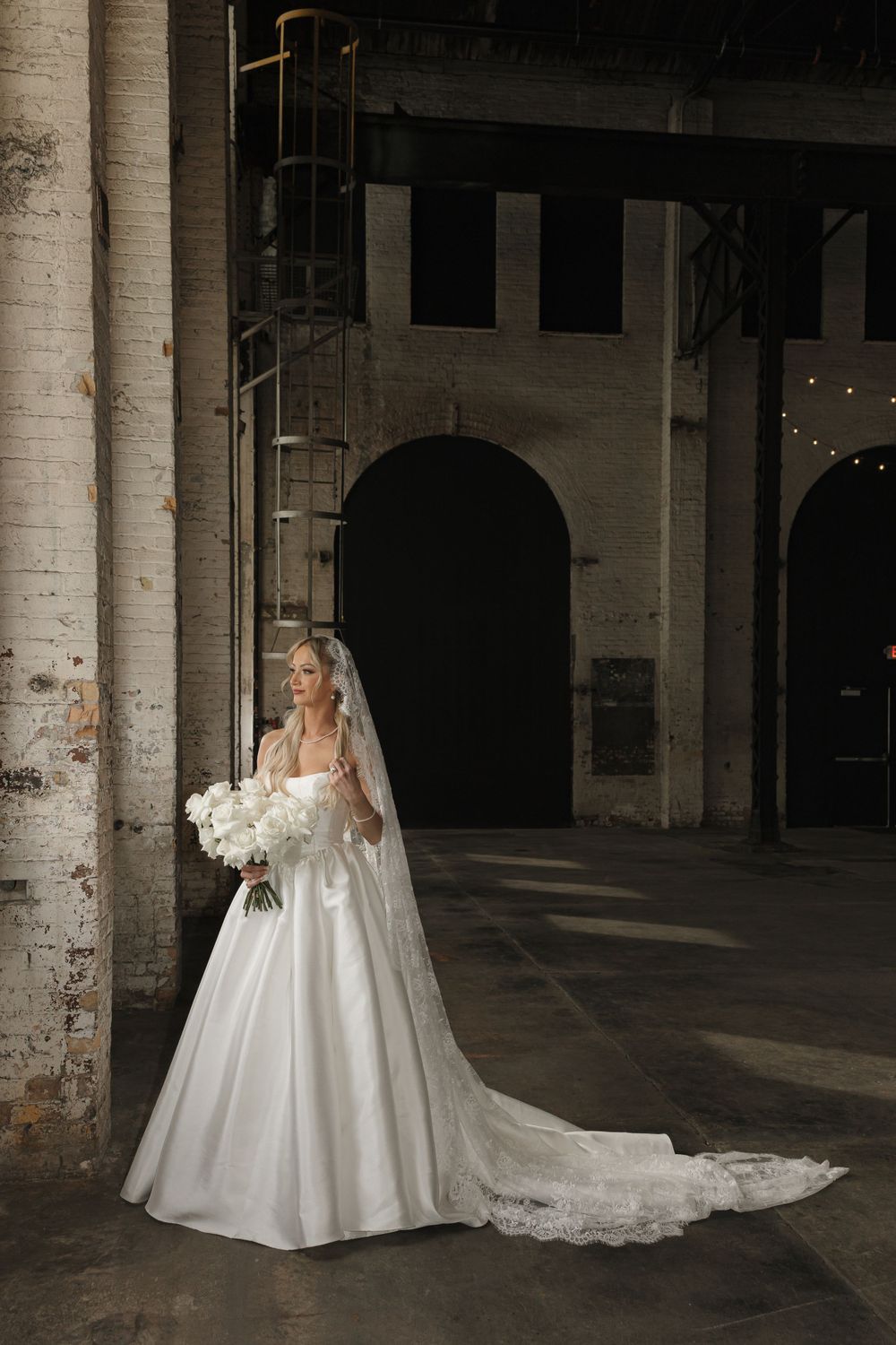 A bride in a classic white ballgown and veil poses in an industrial brick venue.
