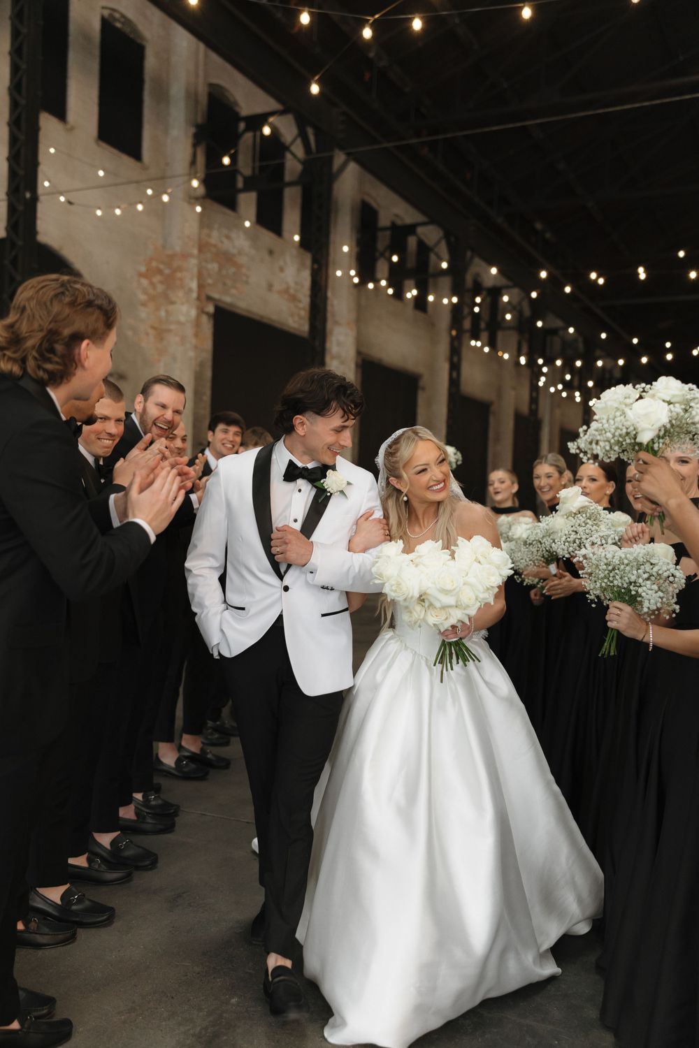 Wedding celebration with guests in black attire applauding the newlyweds under string lights.