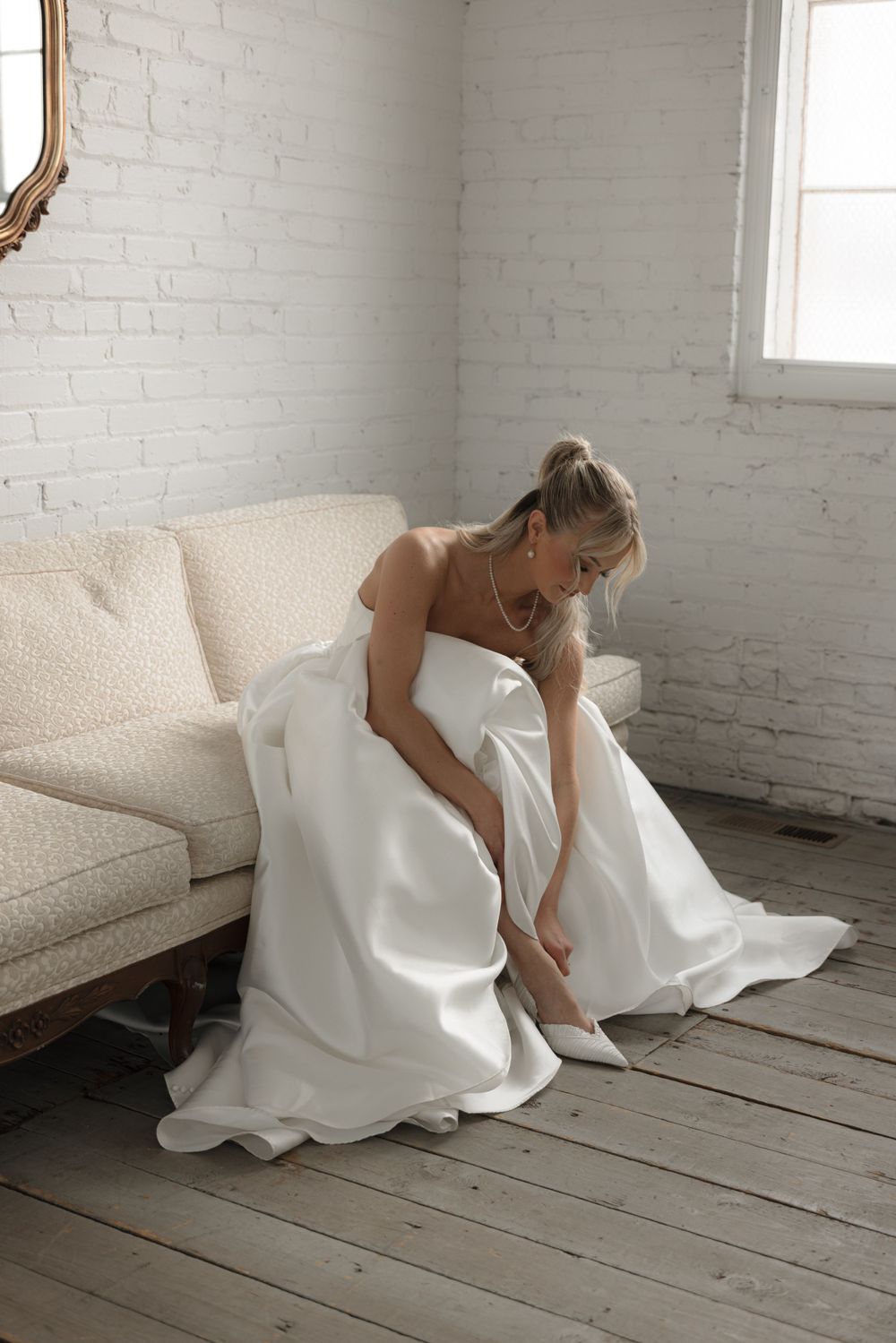 A bride in white wedding dress sits on cream couch putting on shoes in bright room.