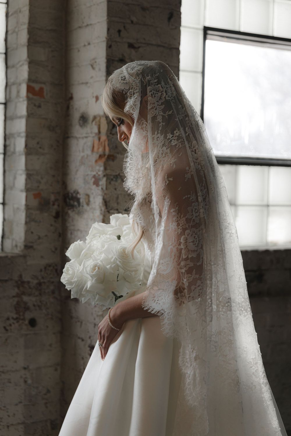 Bride in white dress and lace veil holding white bouquet against industrial brick wall.
