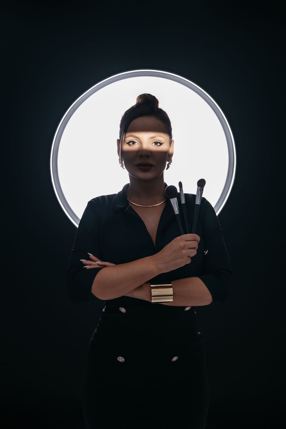 A figure in black attire poses dramatically against a white backlit circular background holding a small object.