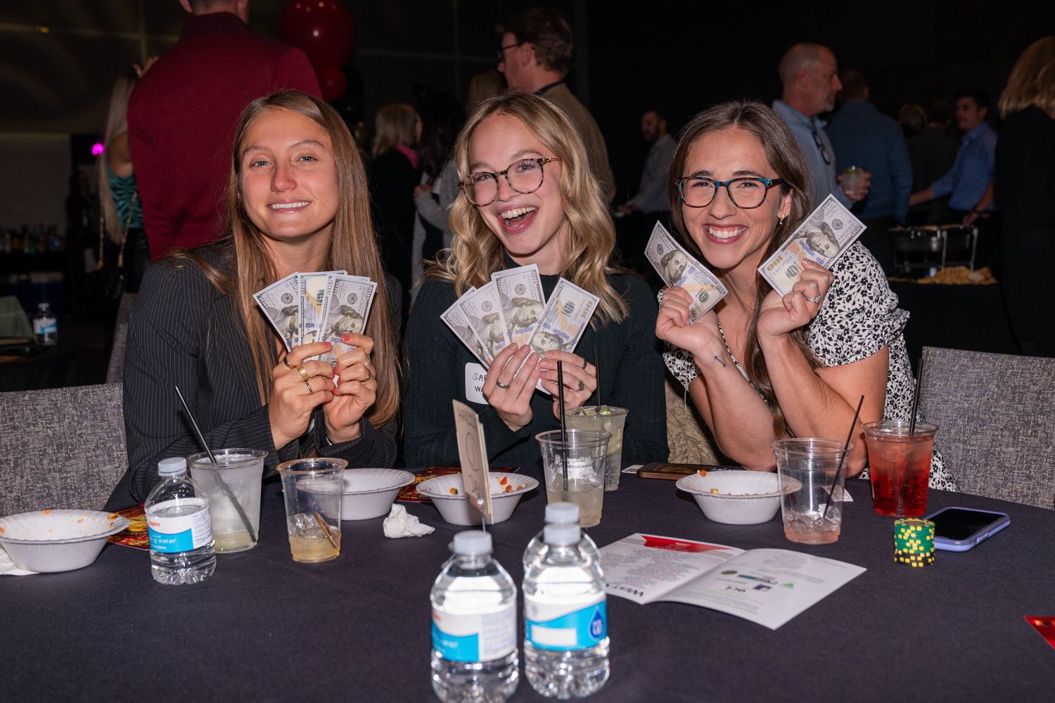 Three people at a table showing playing cards and drinks during a game night.