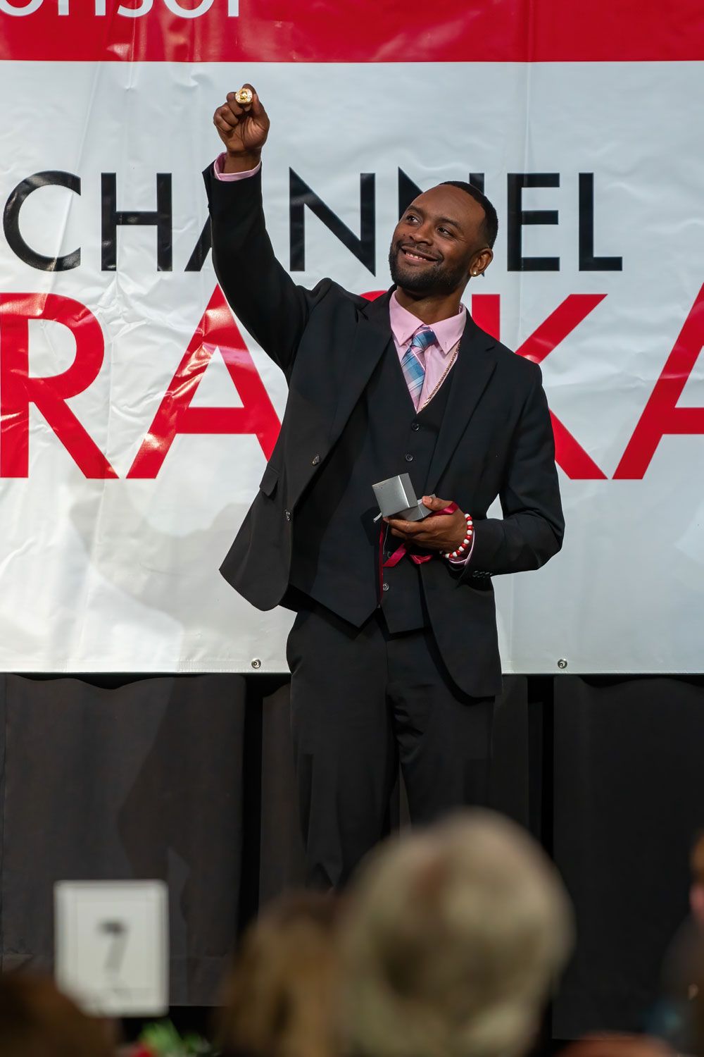 Speaker on stage wearing dark suit raises fist in triumphant gesture at event.