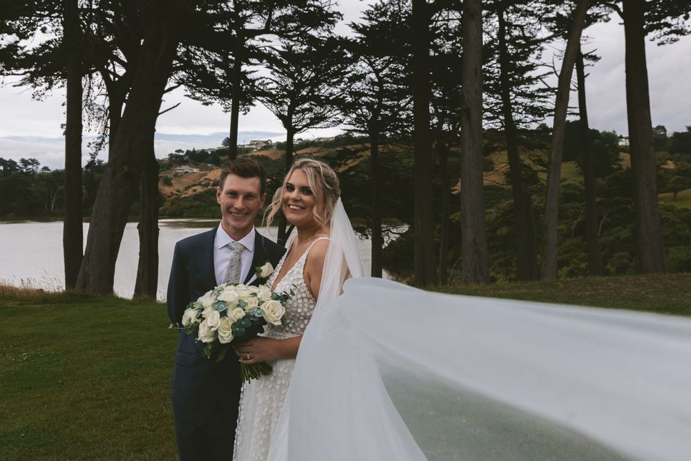 Romantic wedding portrait with couple holding bouquet against lakeside background.