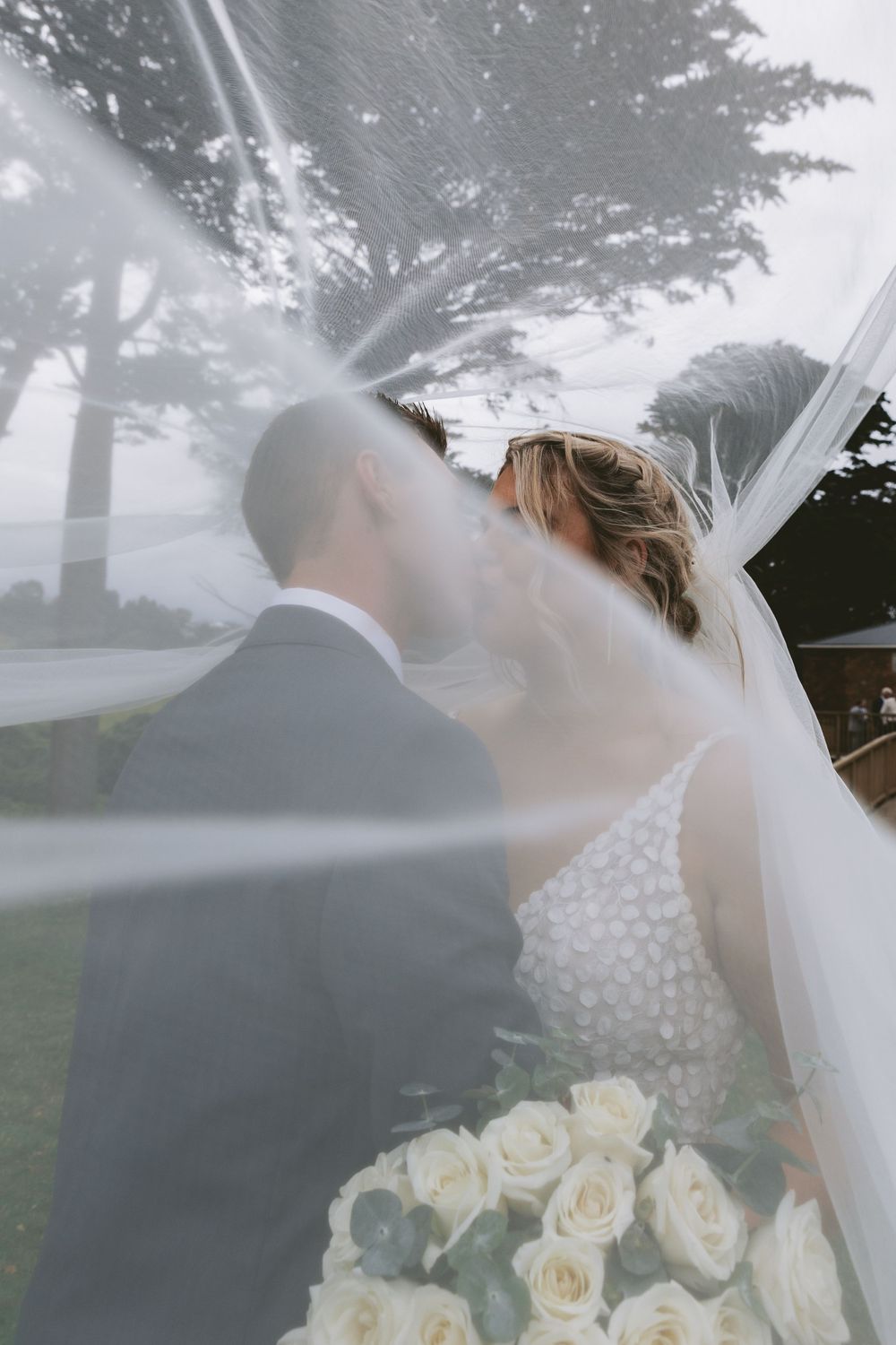 Artistic wedding photo with veil movement and dreamy light filtering through trees.