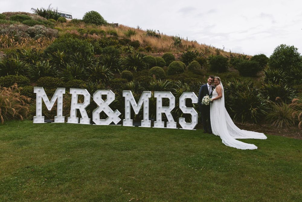 Couple poses by large MR&MRS letters on grassy hillside during wedding celebration.