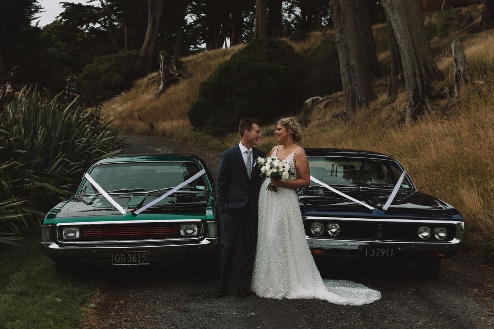 Wedding couple posed between two classic vintage cars on a grassy hillside.