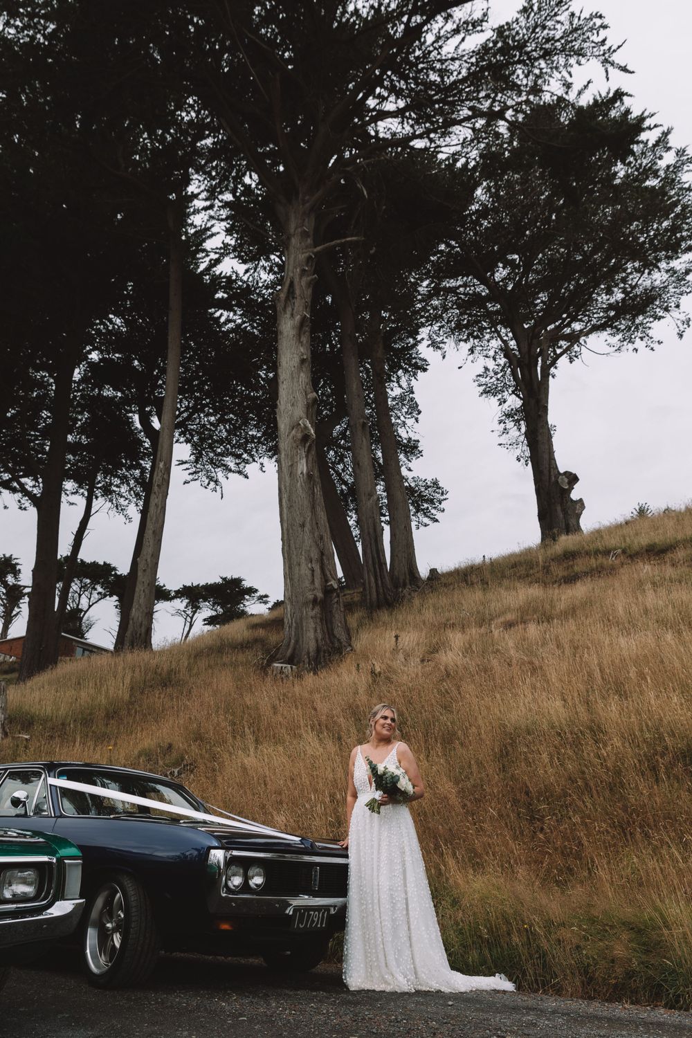 Elegant wedding photography with classic car against towering trees on a rustic hillside.