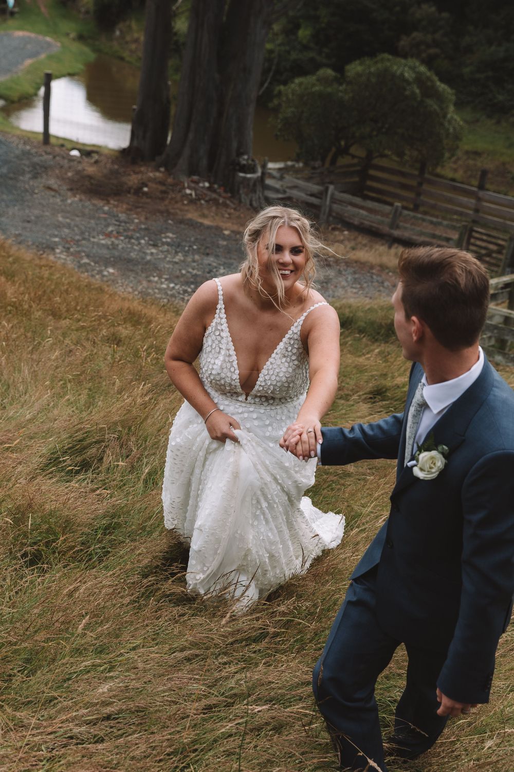 A couple on their wedding day walks through a grassy hillside near a pond, with the bride wearing a sparkly white gown.