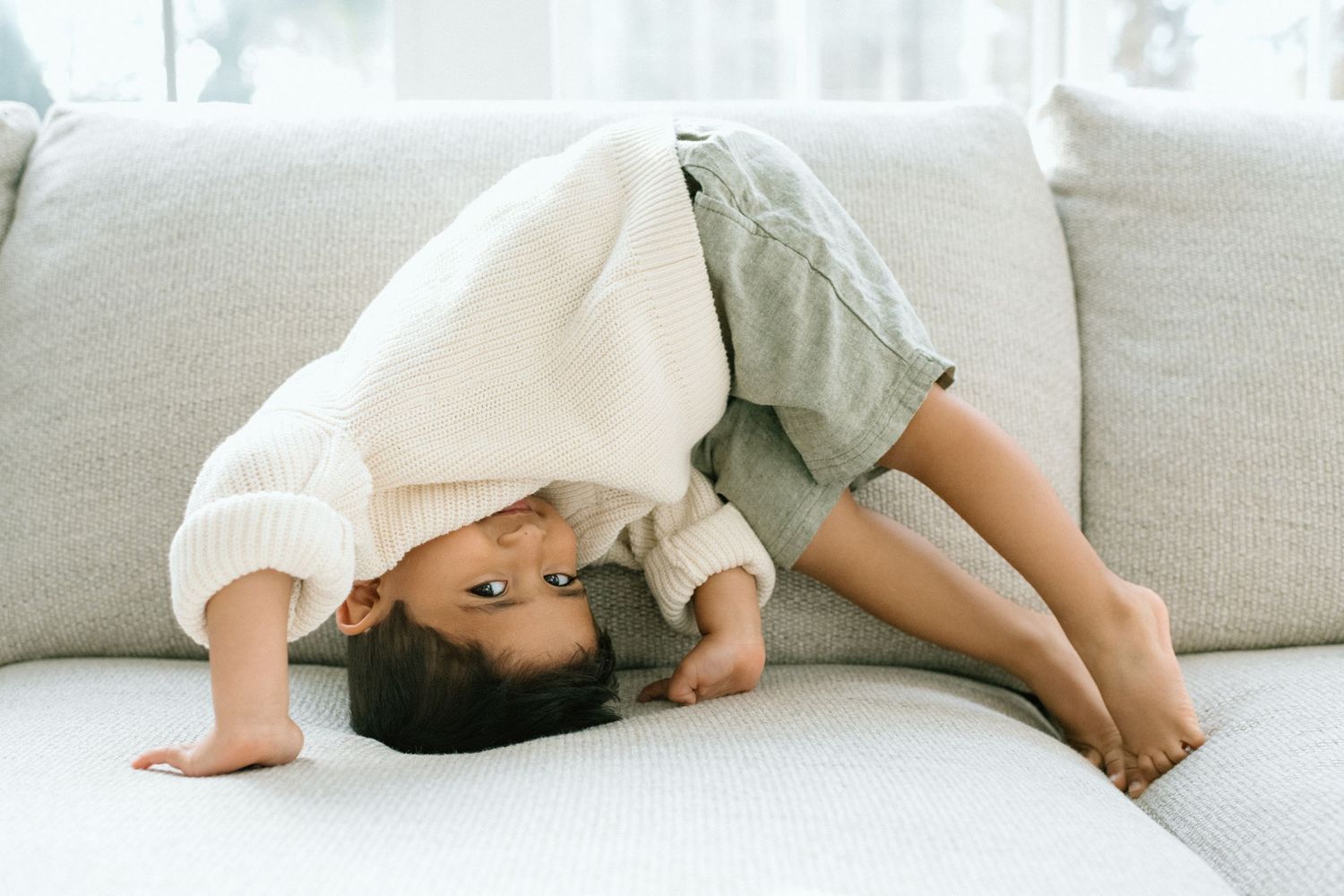 Young child in white shirt playfully bending over backwards on a light gray couch.