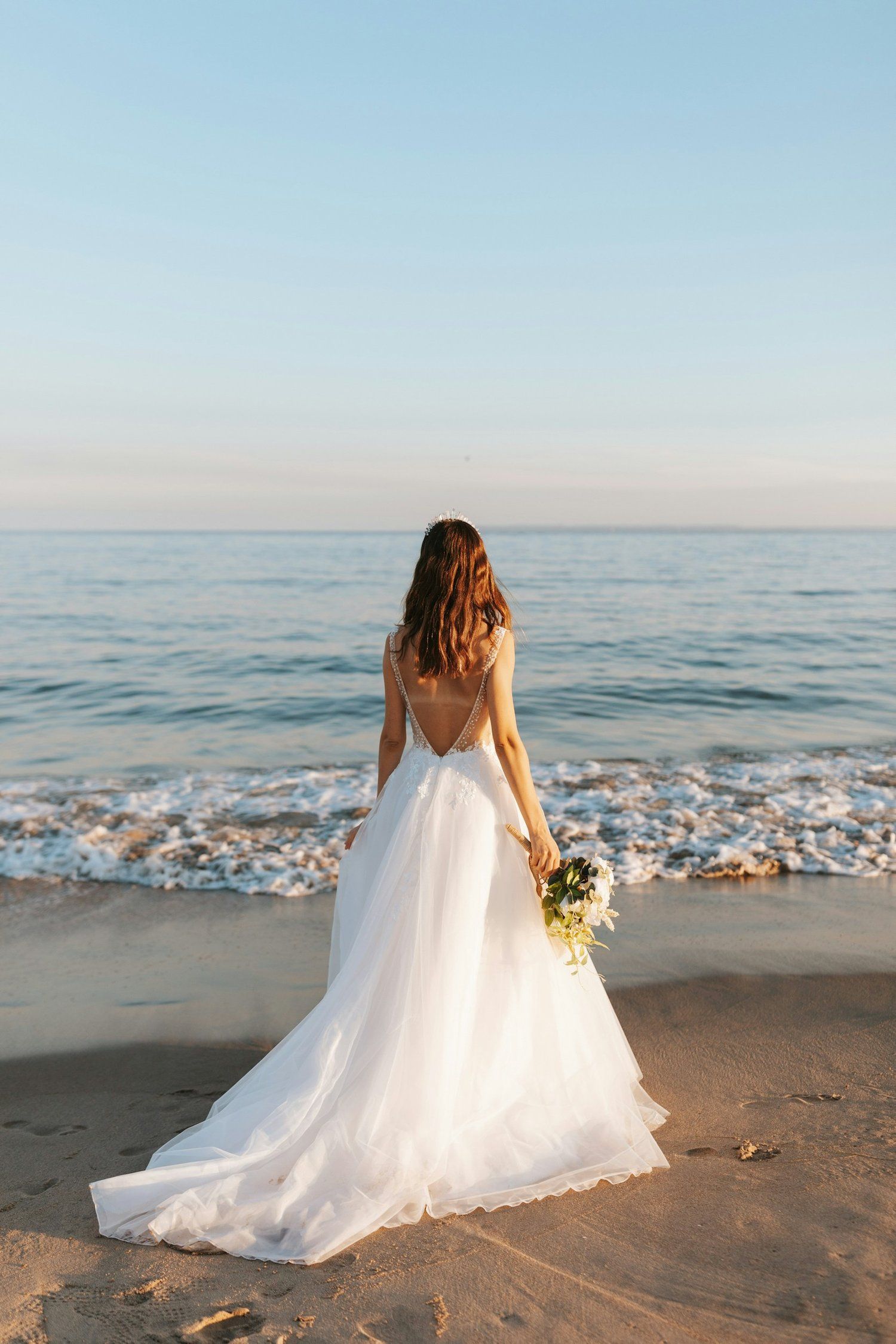 A person in a flowing white wedding dress holds a bouquet while standing on the beach at sunset.