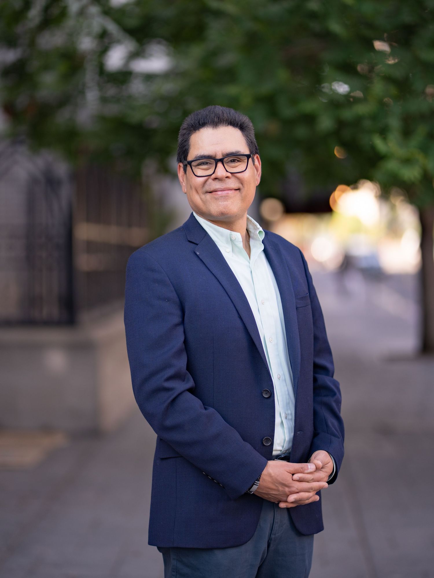 Professional wearing a navy suit and white shirt stands by an iron fence with lush green shrubs in the background.