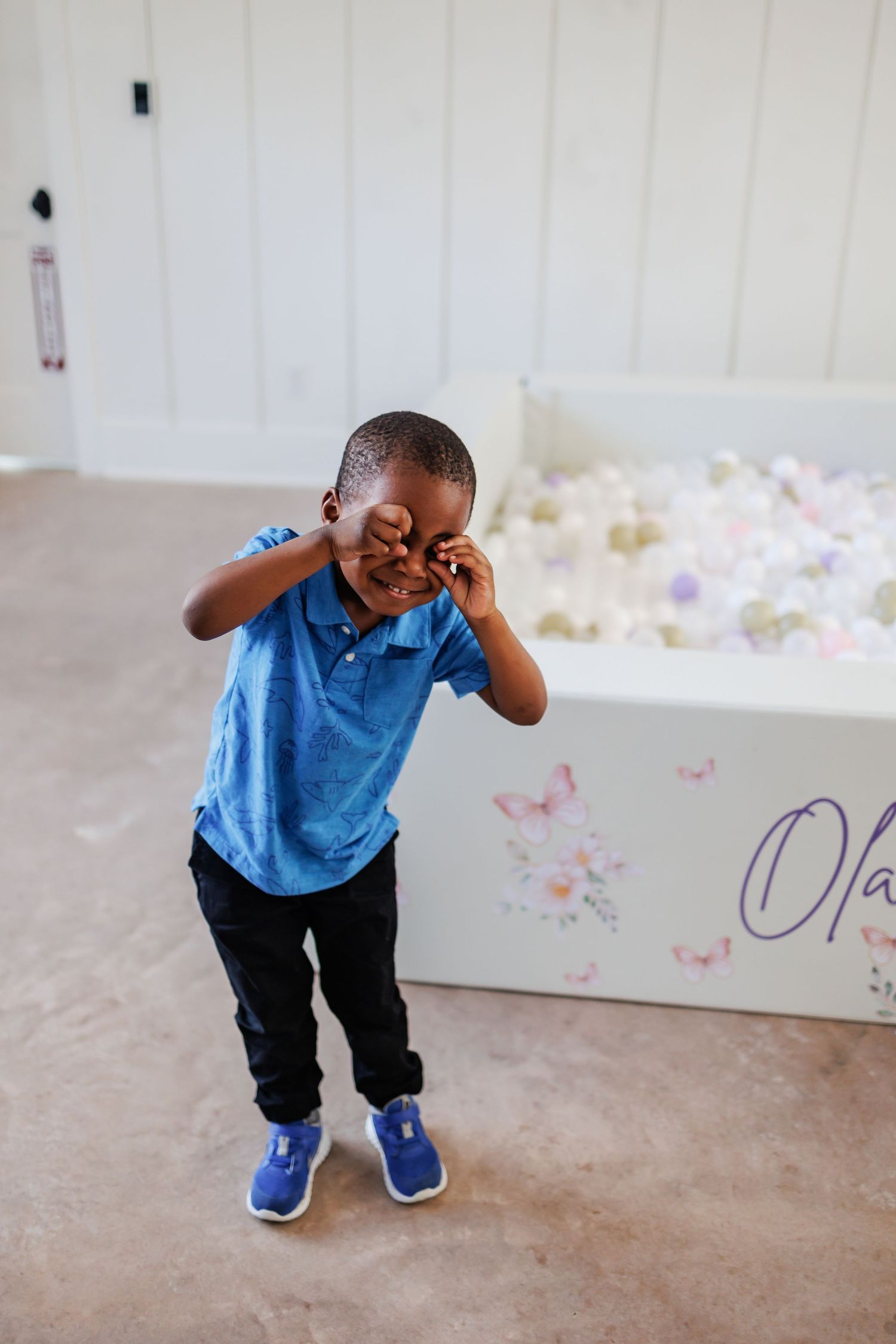 Young child wearing a blue polo shirt and dark pants smiles in front of a white ball pit at a first birthday party