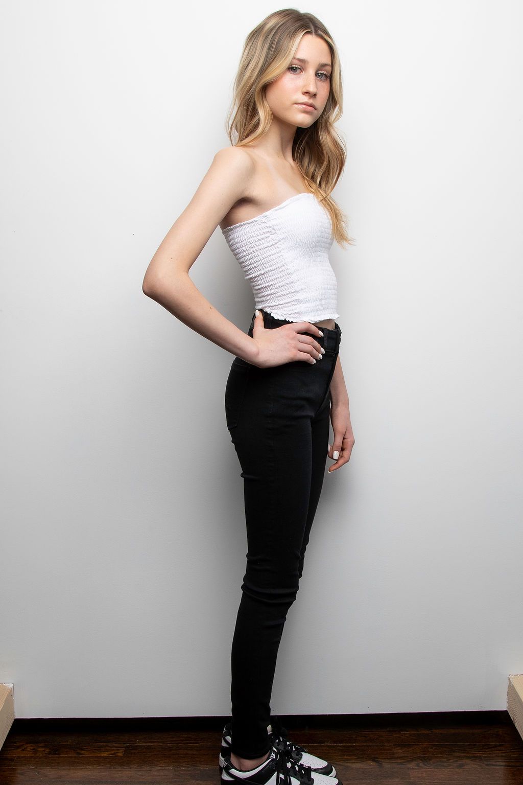 A person in a casual outfit with a white strapless top and black skinny jeans poses against a white wall.