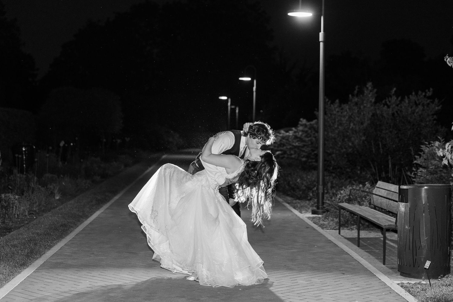 A couple in wedding attire share a romantic moment under streetlights on a path at night in black and white.