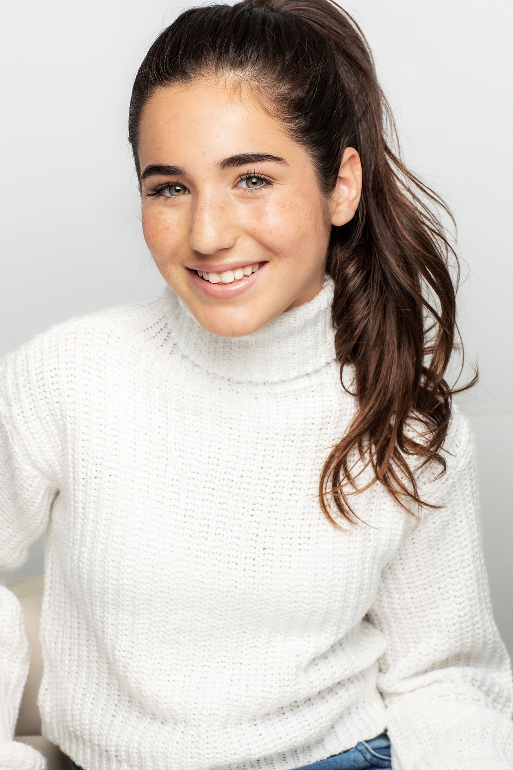 A smiling figure with dark hair in a ponytail wearing a white turtleneck sweater against a light background.
