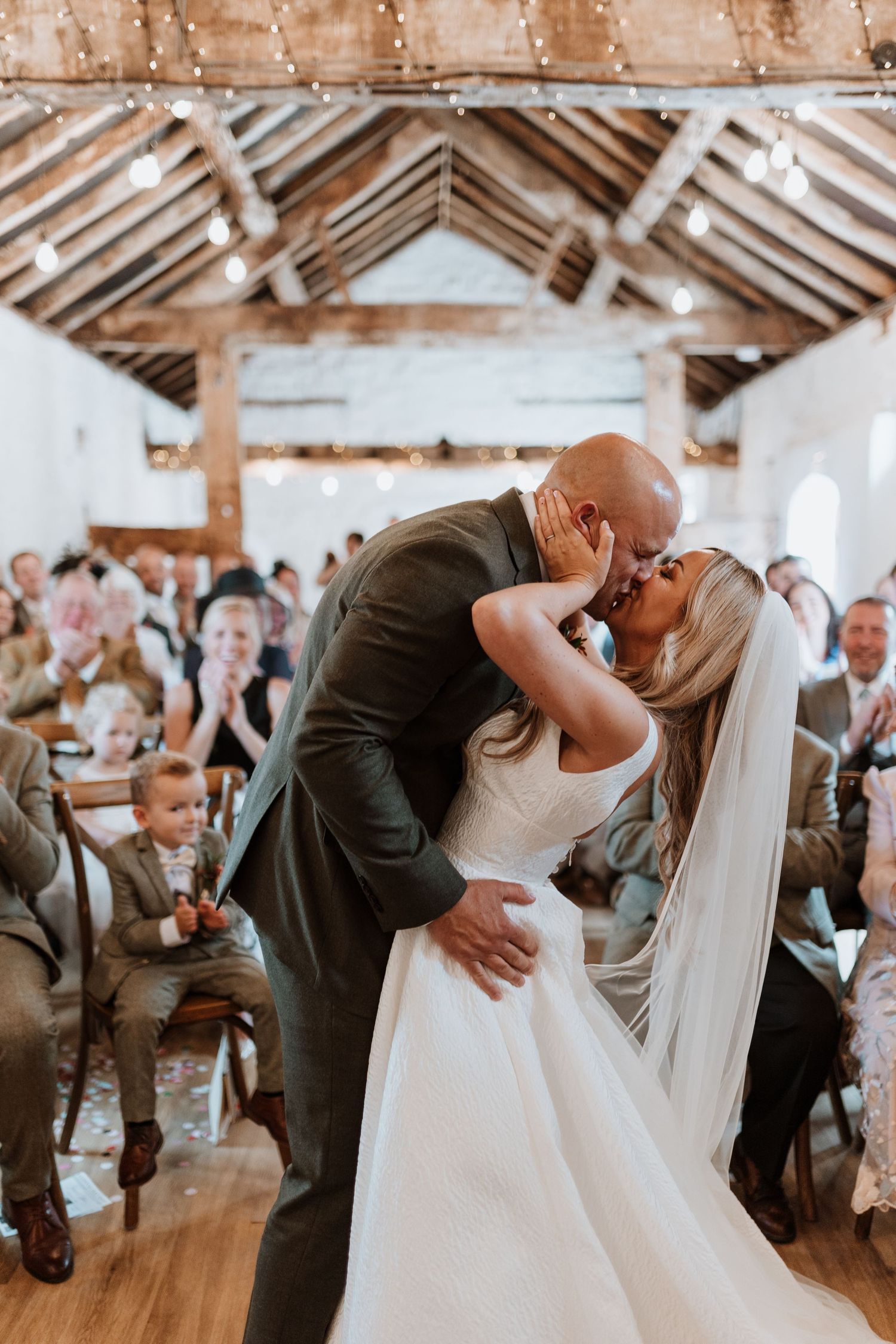 A bride and groom share their first kiss as a married couple in a rustic wooden barn venue surrounded by wedding guests at Stanford Farm.