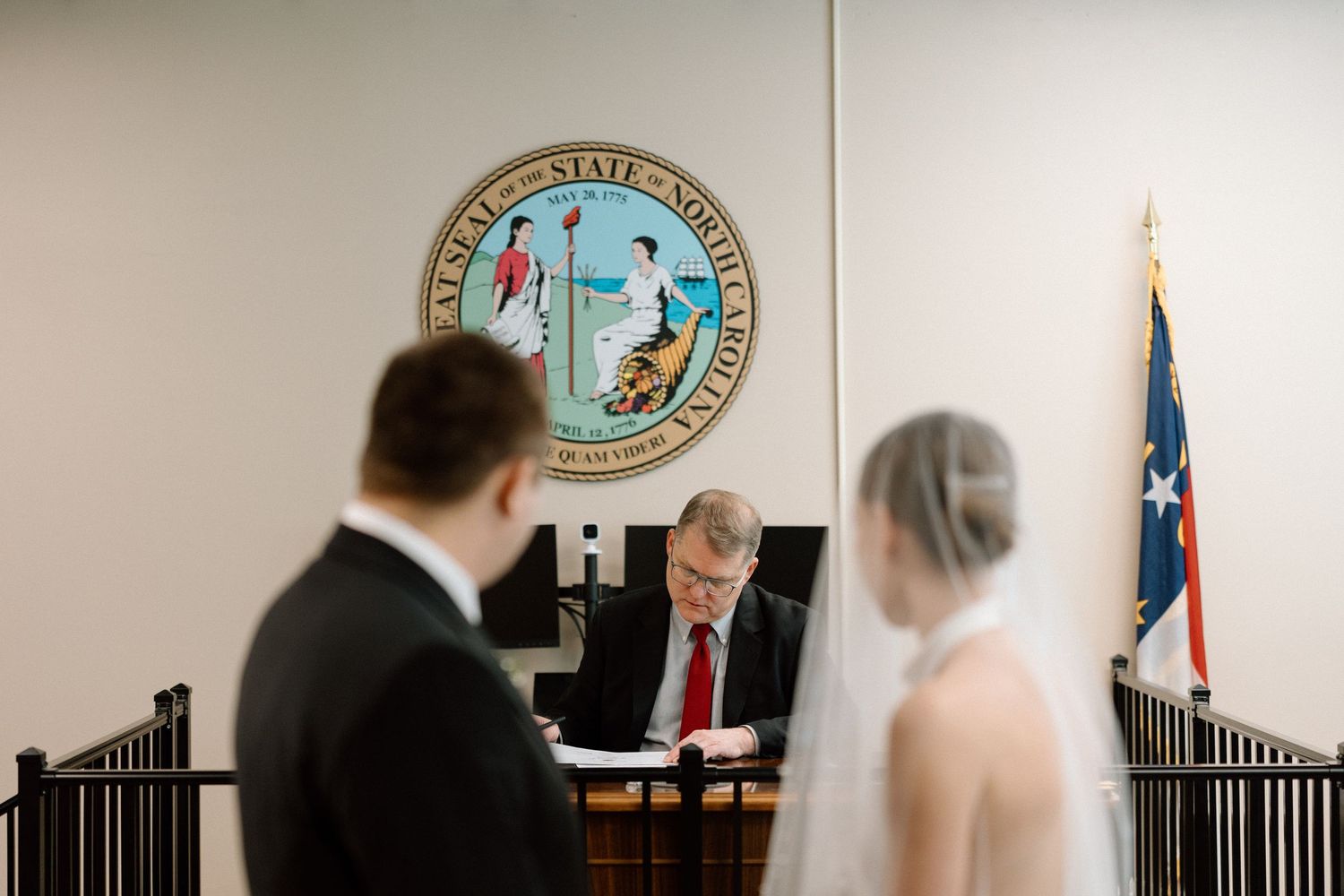 A civil ceremony takes place in a courthouse office with a North Carolina state seal visible on the wall.