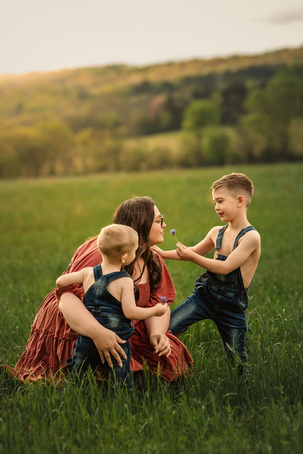 A mother enjoys a moment with her young boys in a lush green grassy field at sunset.