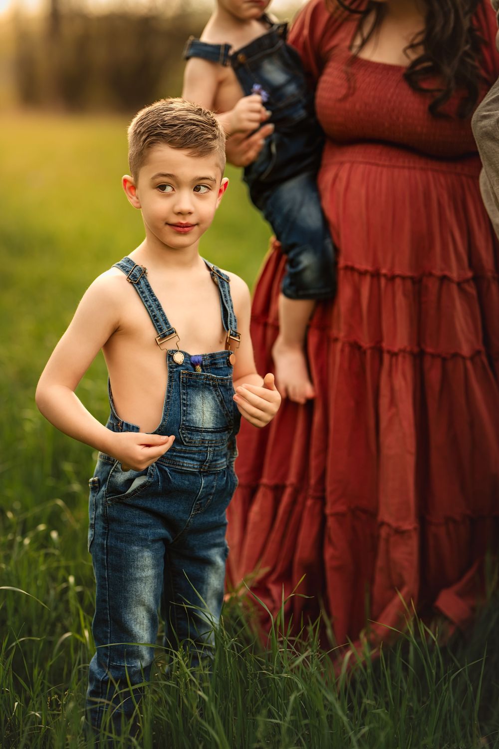 Young child in blue denim overalls stands in a grassy field next to his mother as the sun sets behind them.