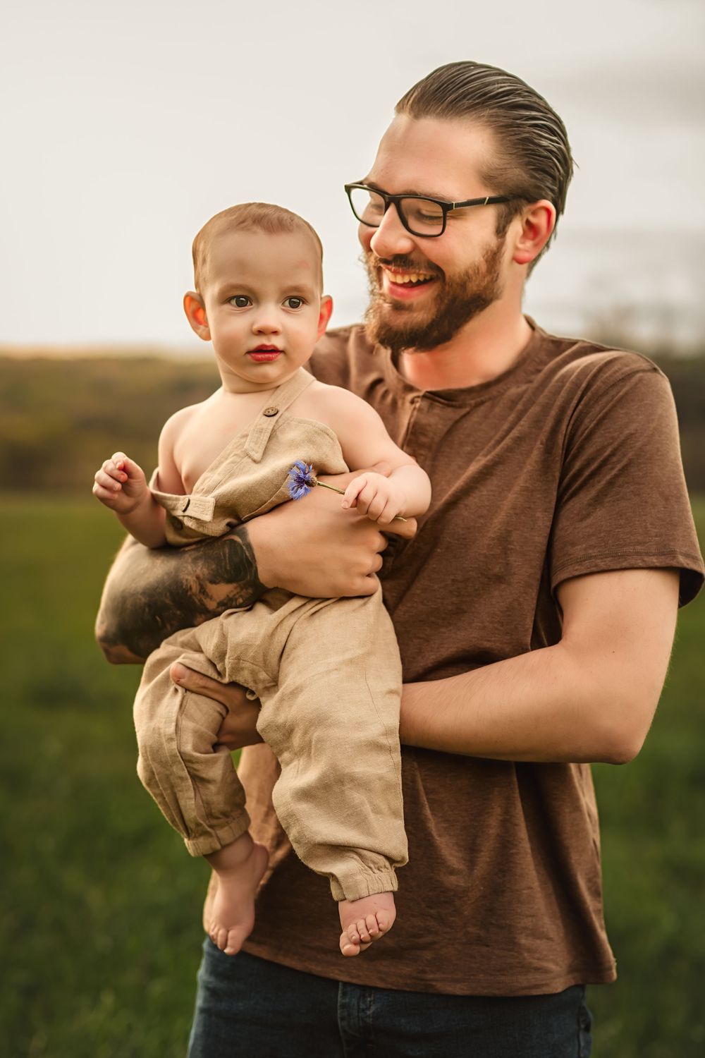 A dad wearing glasses and a brown t-shirt holds a small child in beige clothing on a grassy hillside at sunset.