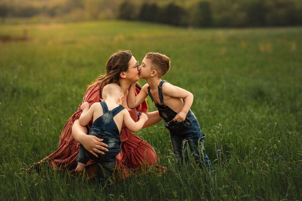 A mother embraces 2 sons affectionately in a lush green meadow during a warm summer day.