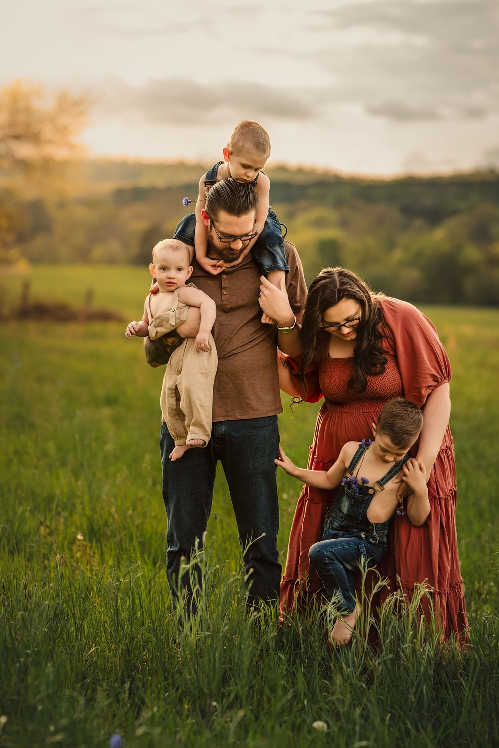 A mom and dad enjoy a tender moment with their 3 sons in a sunlit meadow during golden hour as the sun sets over rolling hills.