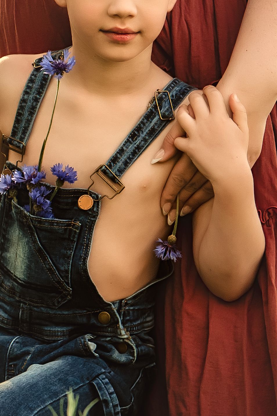 Artistic photo showing a young boy in overalls with blue cornflowers tucked into pockets against his mother's burgundy dress.