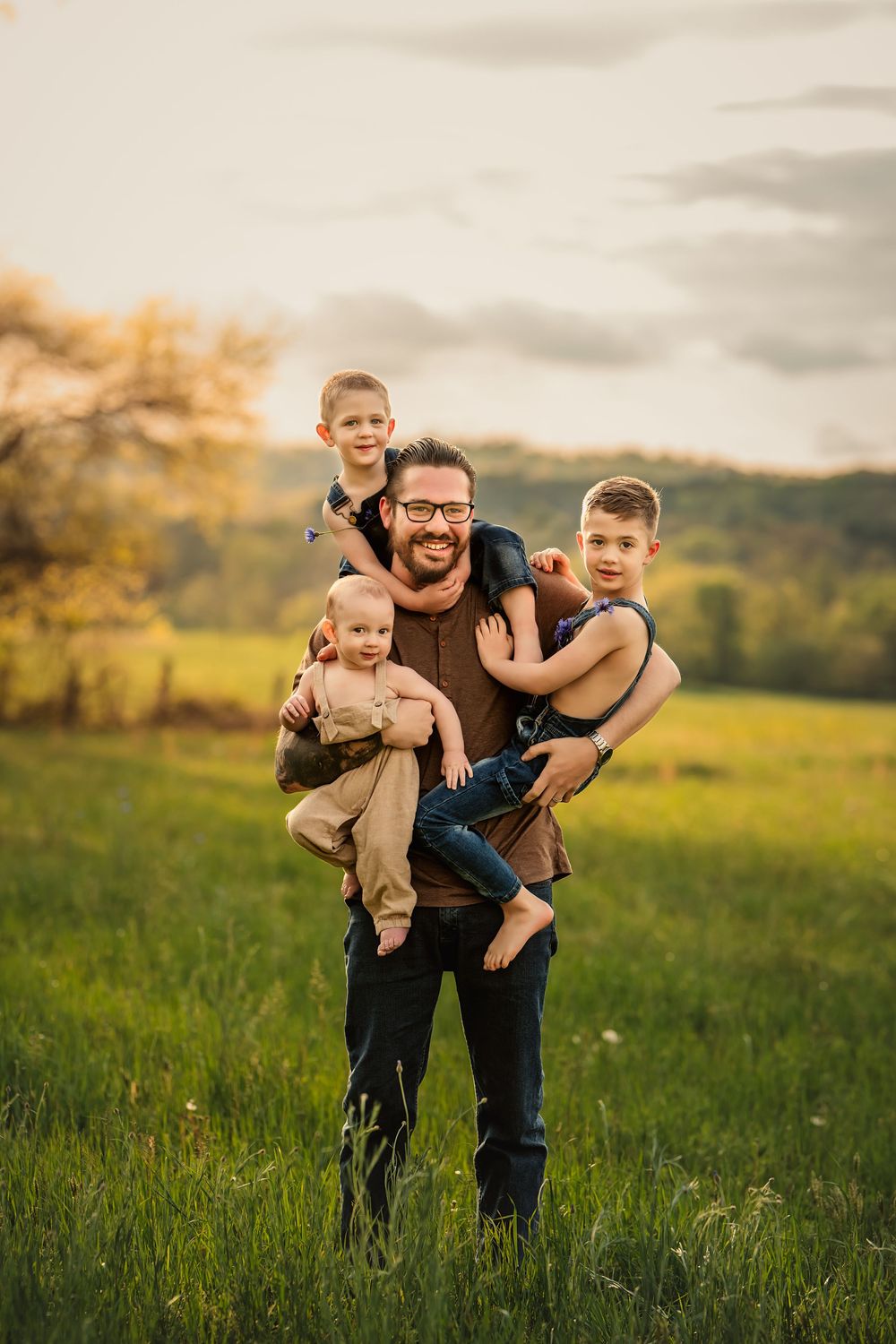 A dad stands in a grassy meadow at sunset holding all three children at once in a playful pose.