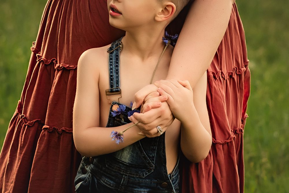 A young child in denim overalls holds onto his mother's arm while holding a purple wildflower.