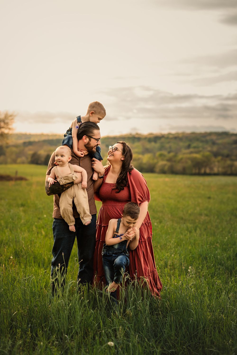 Mom, Dad, and 3 boys embrace in a green meadow at sunset against rolling hills in warm lighting for family photo session.