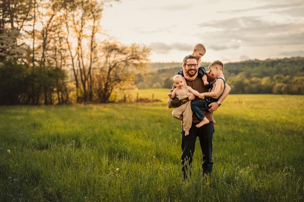 A dad gives a playful piggyback ride to his 3 sons in a grassy field at sunset with trees silhouetted in the background.
