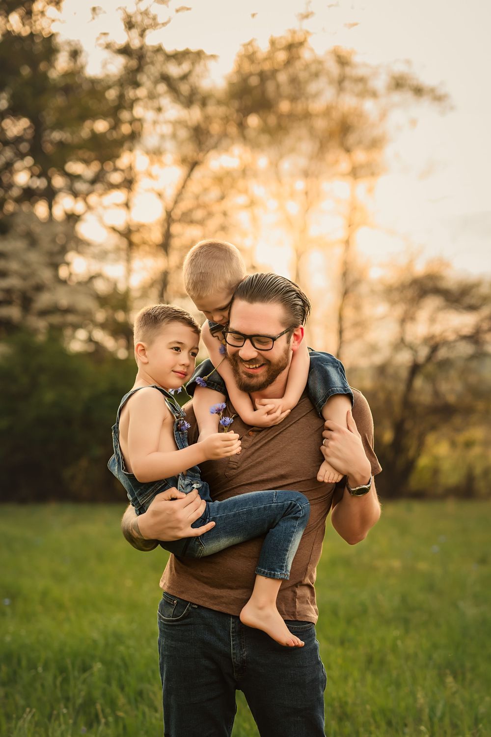 A playful moment captured at sunset in a grassy field as dad tries to hold 2 young boys at the same time.