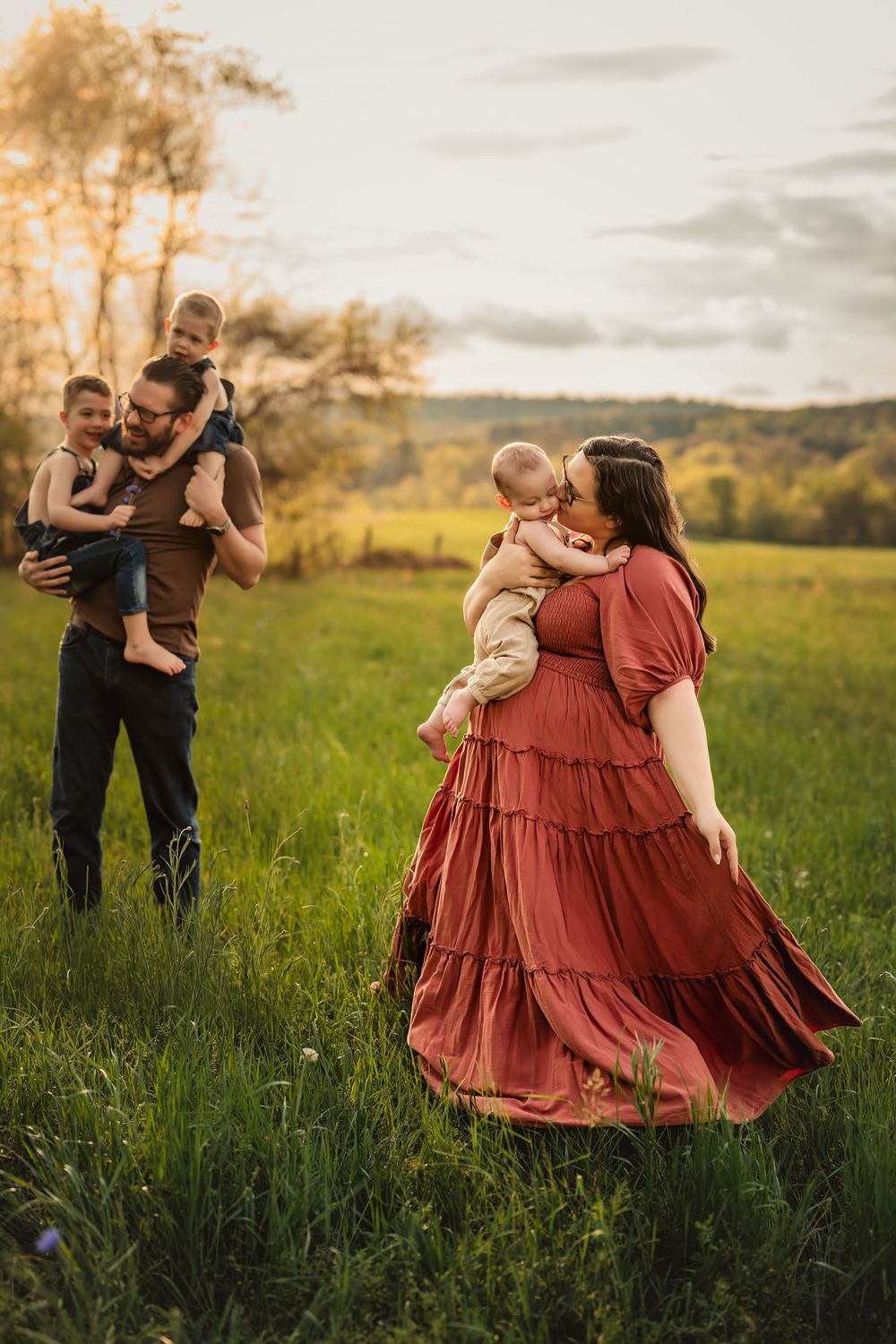 Candid family moments captured in a grassy field at sunset with warm golden light. Dad plays with older boys while mom dances with baby.