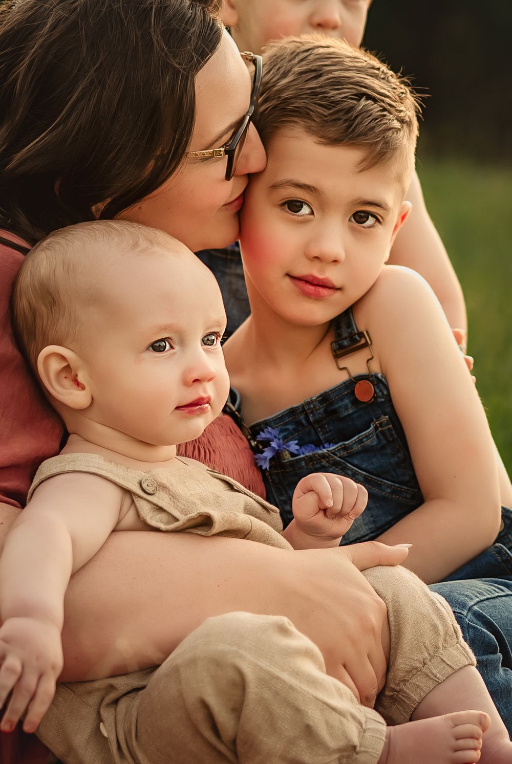 Loving family portrait captured outdoors showing mom in glasses snuggling close with two young children.