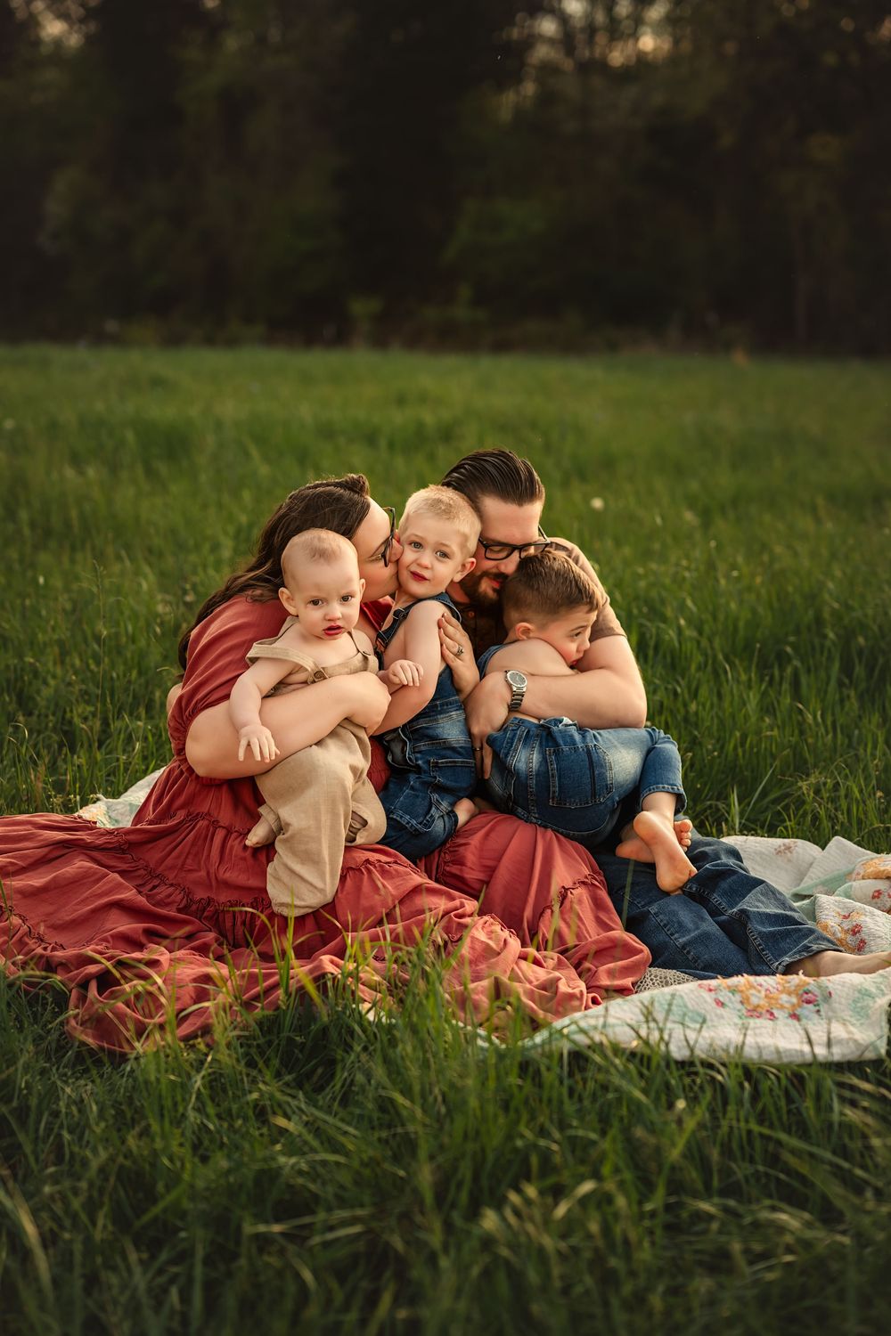 A loving family cuddles together on a blanket in a green field during golden hour.