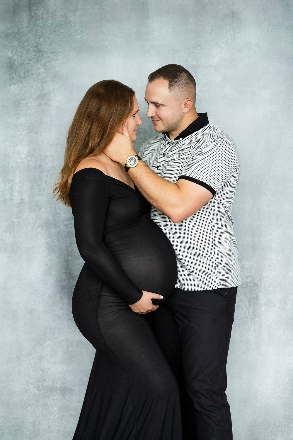 Intimate maternity photo of couple in black dress and gray shirt against textured gray background.