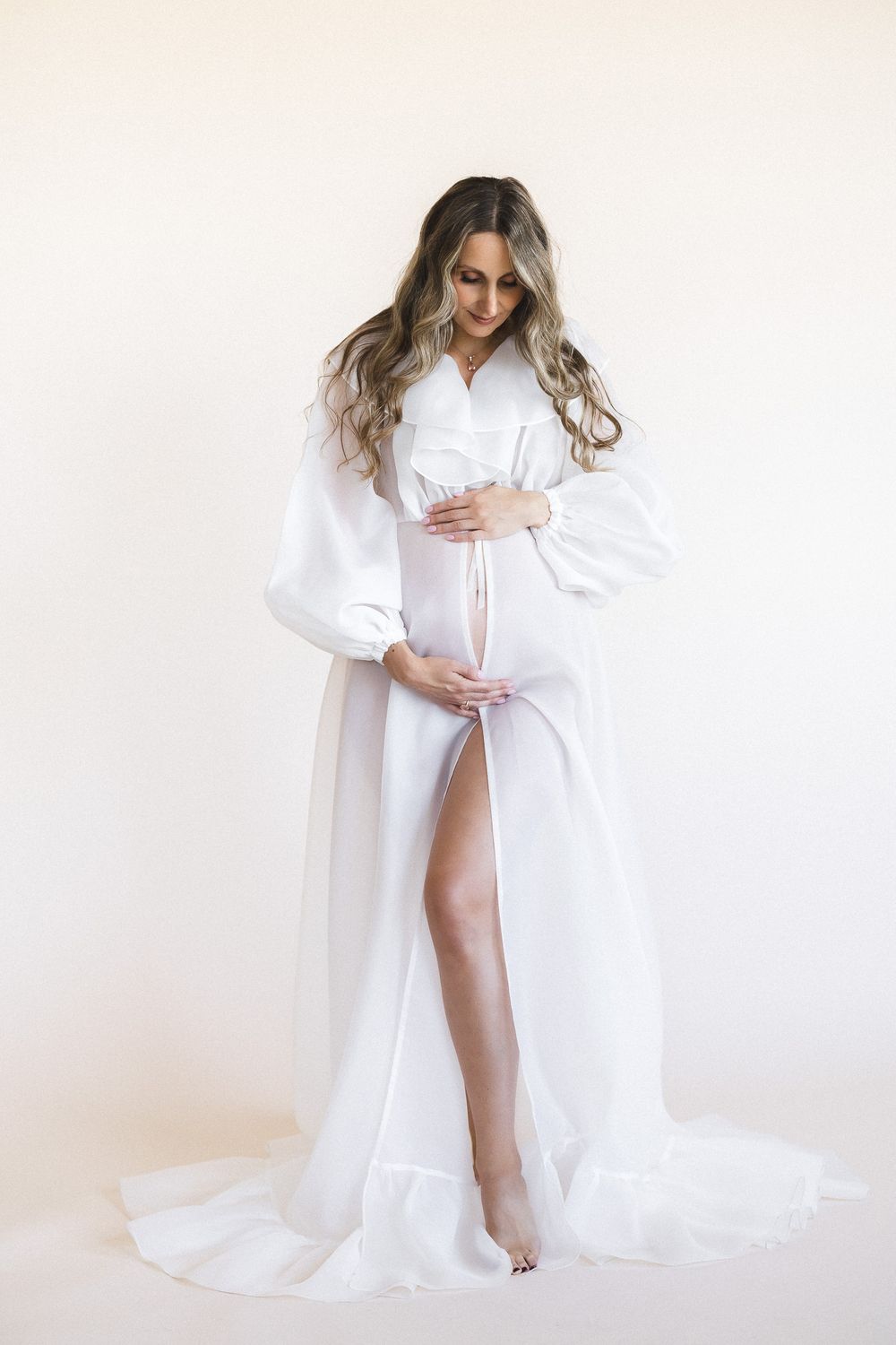 Elegant maternity portrait in flowing white robe against light background.