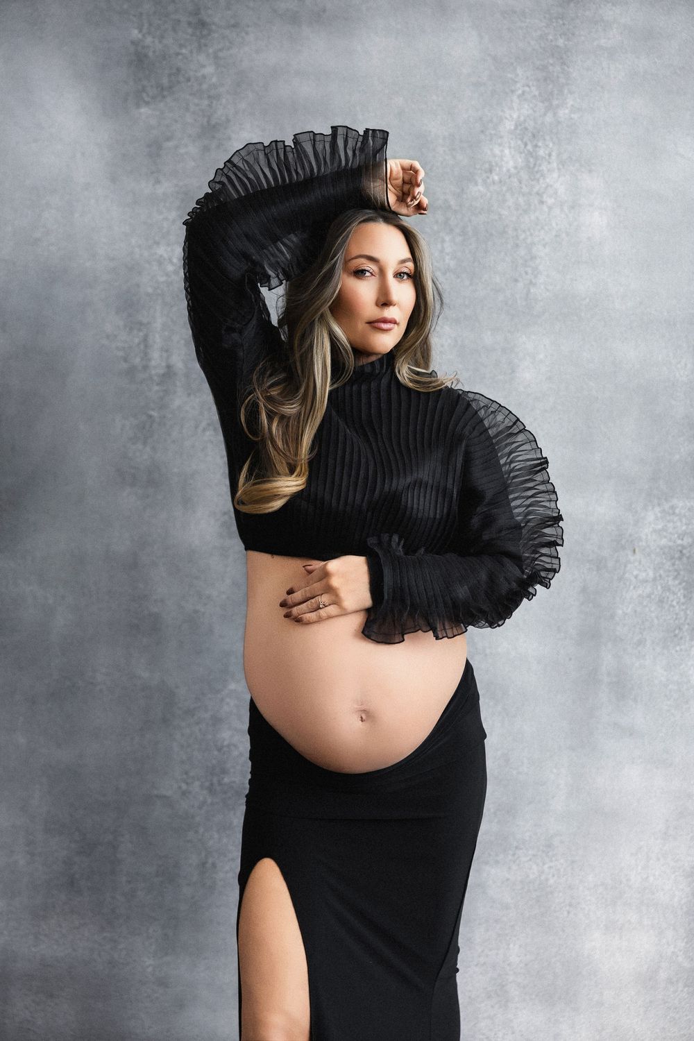 Artistic maternity photo with dramatic ruffled sleeves and exposed belly against textured grey background.