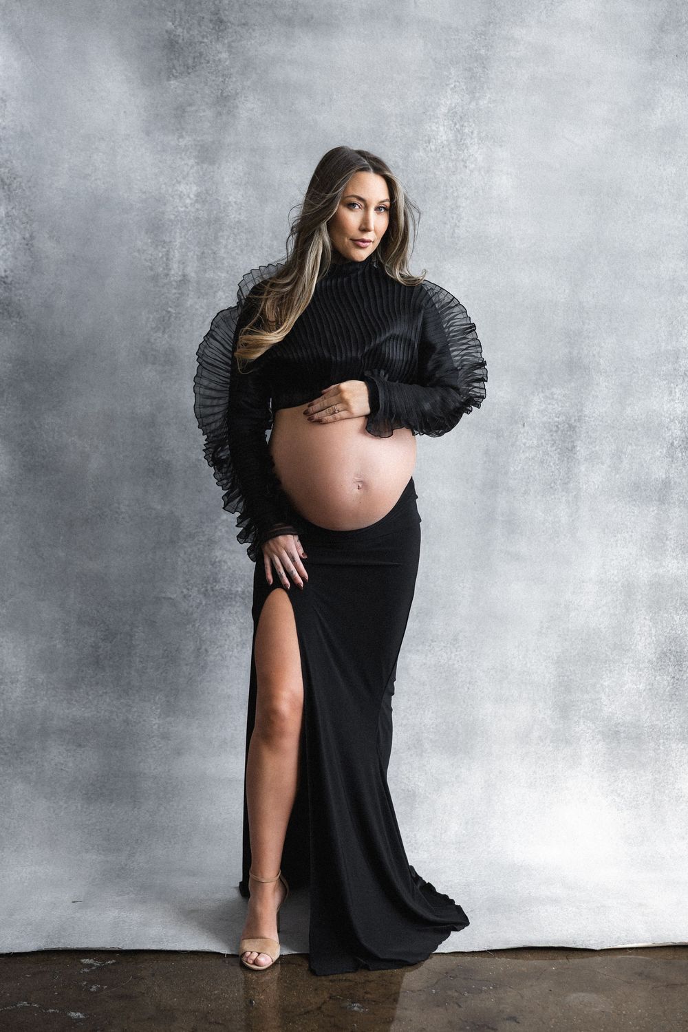Dramatic maternity photo featuring black gown with ruffled sleeves and high slit against grey backdrop.