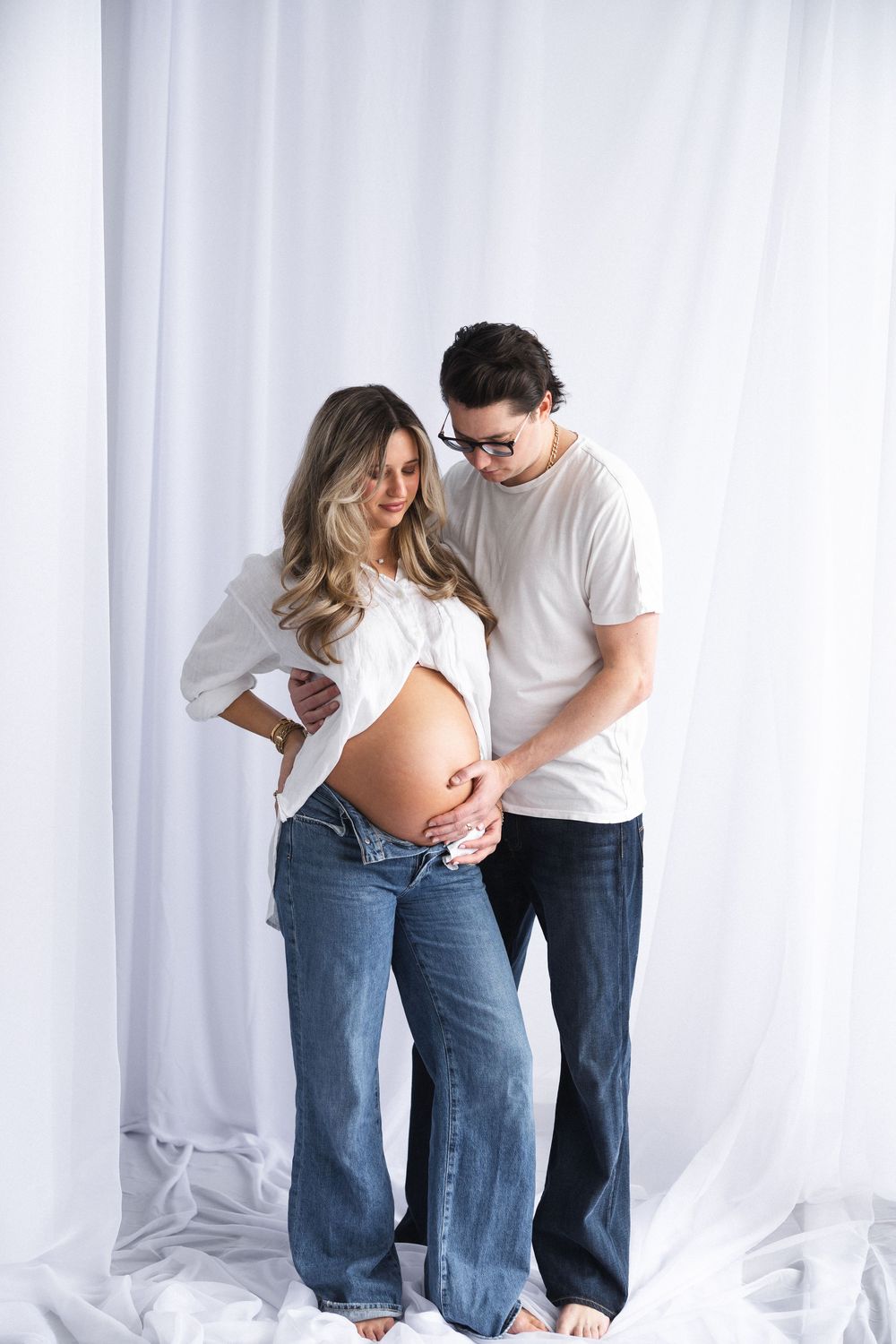 Intimate couple's maternity shoot in casual white and denim against white backdrop.