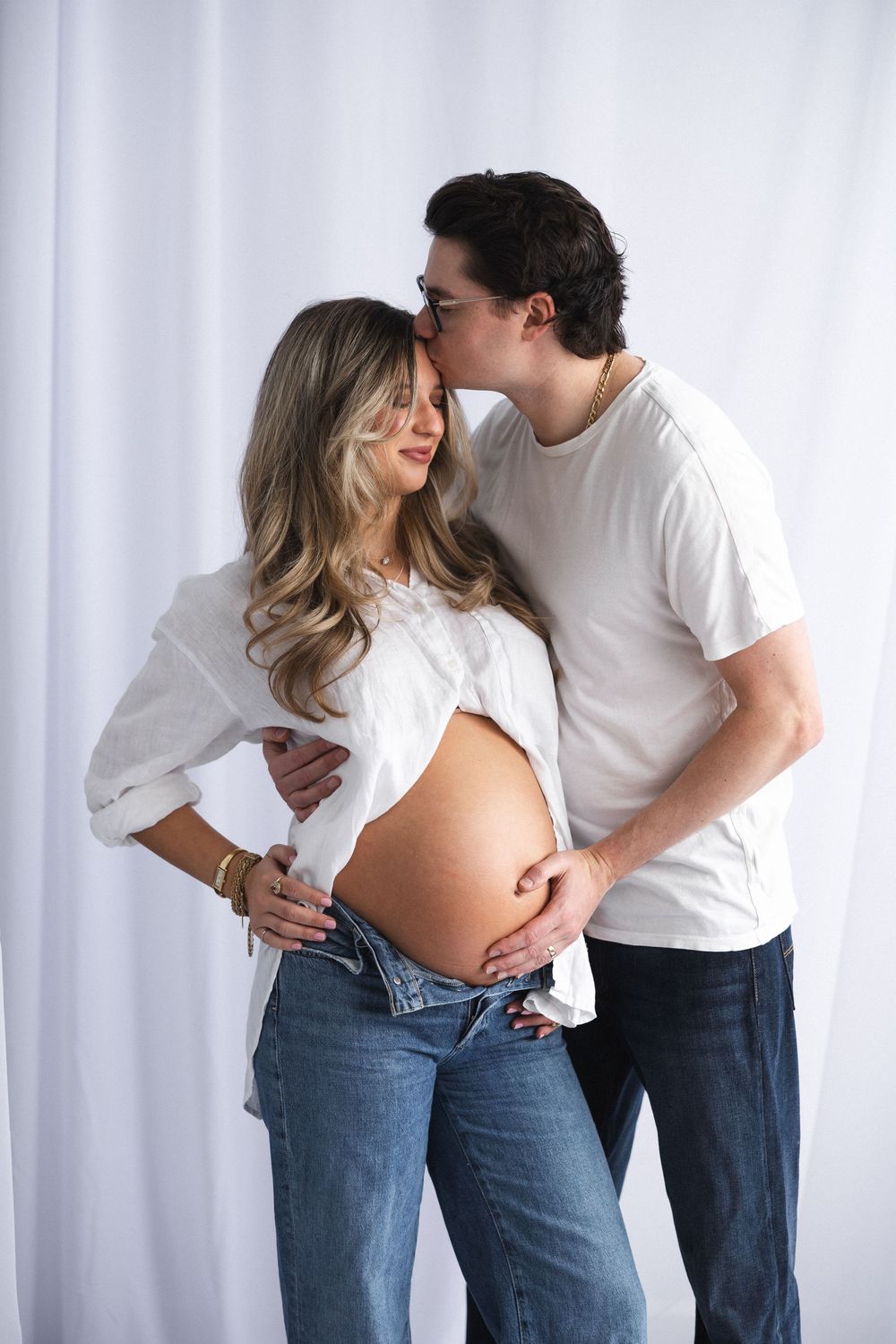 Sweet candid moments between expecting couple in casual wear against white backdrop.
