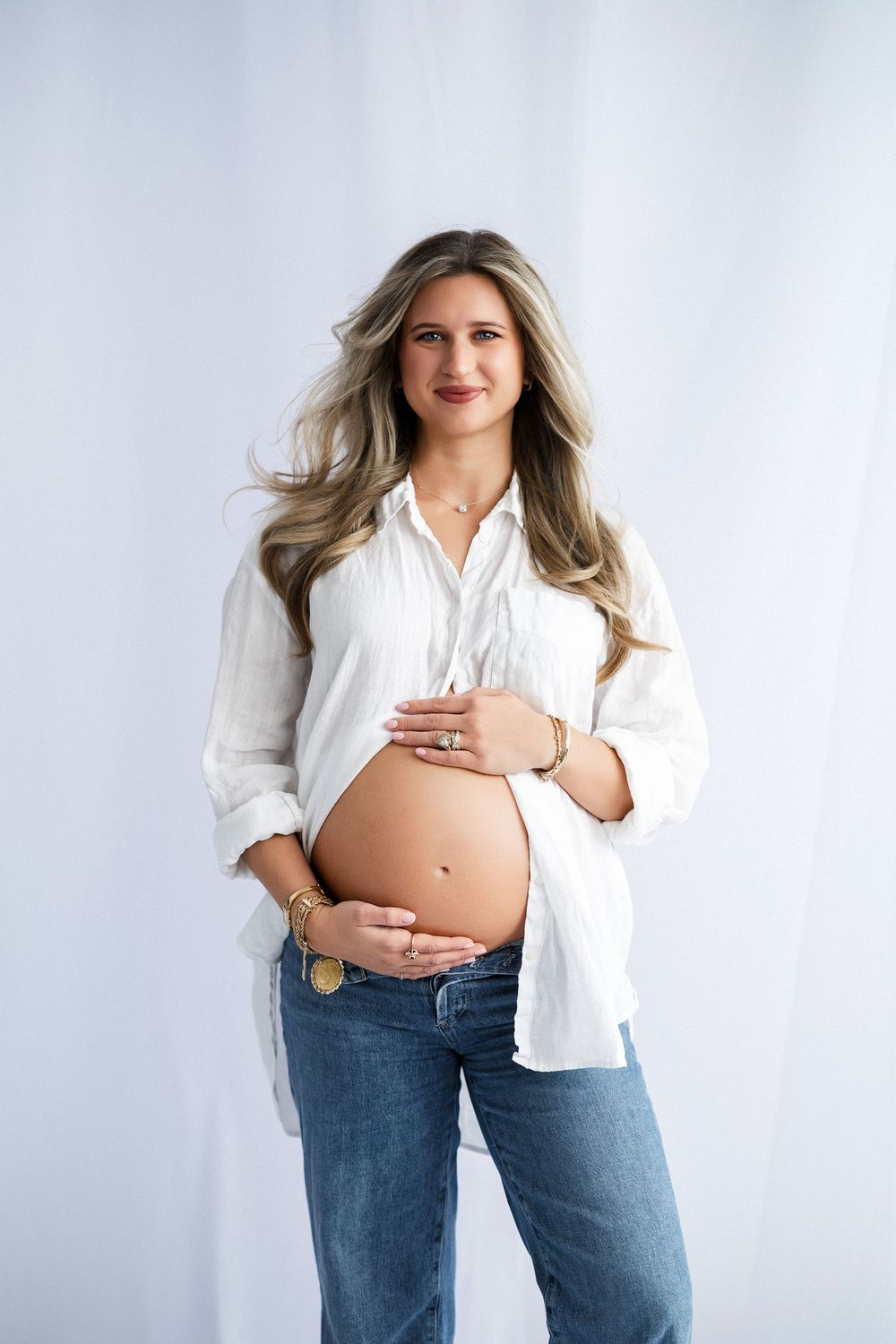 Casual maternity portrait in white button-down shirt and jeans against light backdrop.
