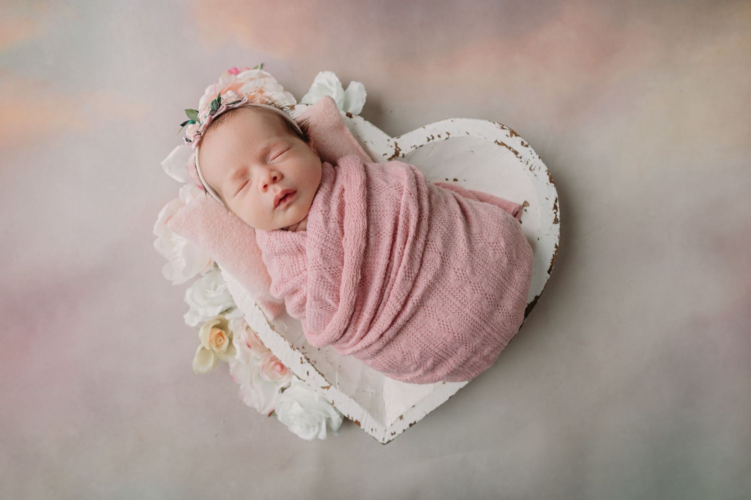 Sleeping newborn wrapped in soft pink blanket nestled in heart-shaped bed with delicate flowers and white fabric during twin baby photoshoot