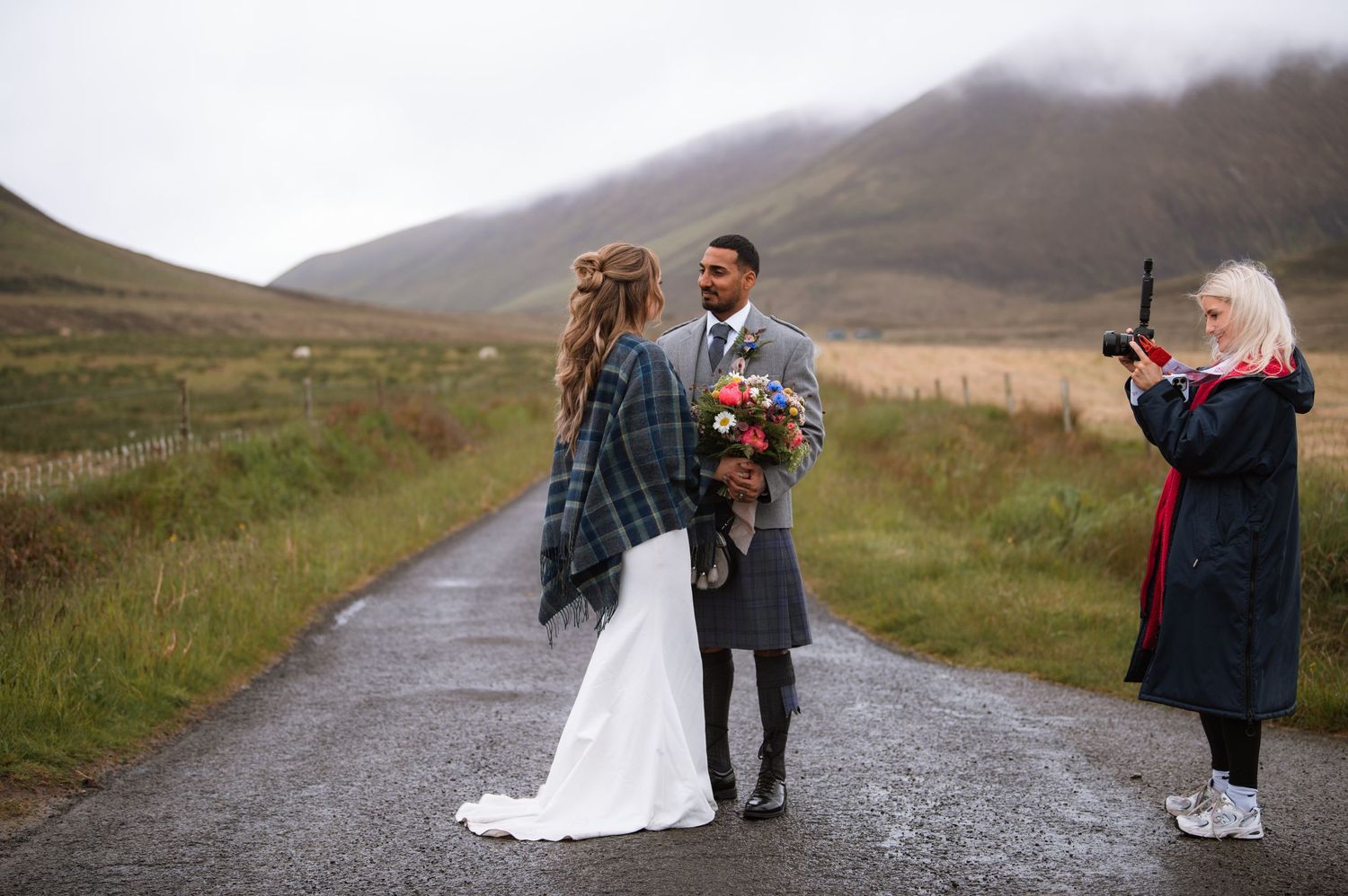 A couple wearing traditional Scottish wedding attire poses on a misty country road in the Scottish Highlands.